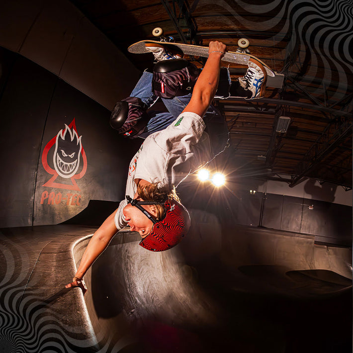 Skateboarder performing a trick in an indoor skate park with the Pro-Tec x Spitfire logo on the wall.
